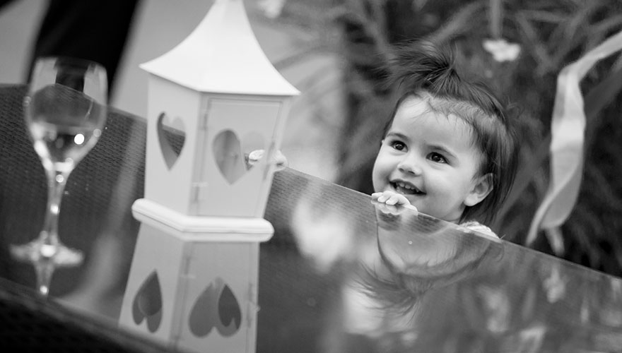 Bei einer Hochzeit steht ein kleines ein bis zweijähriges Mädchen an einem Tisch und lächelt als es eine Laterne mit Herzfenstern erblickt.