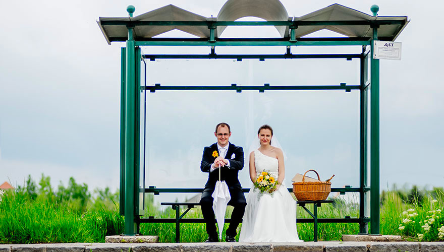 Ein Brautpaar sitzt in einem Bus-Haltestellen-Häuschen. Der Bräutigam hat in der Hand einen Schirm. Neben der Braut steht noch ein Picknick-Korb.
