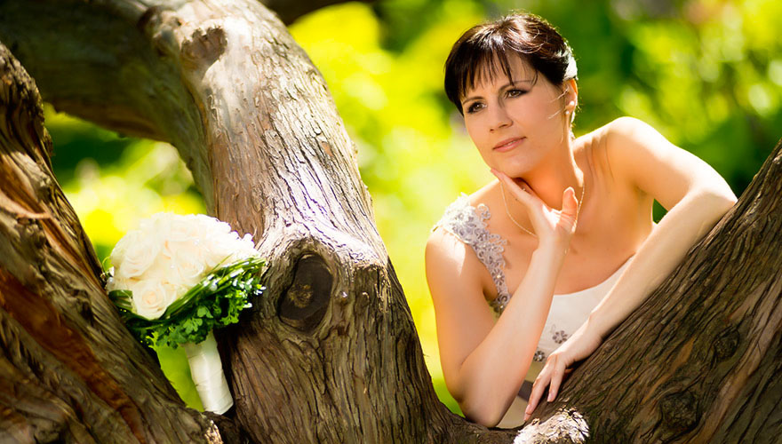 Eine Braut steht hinter einem Baum und hat sich auf einer Astgabelung aufgestützt. Sie sieht erwartungsvoll in die Ferne.