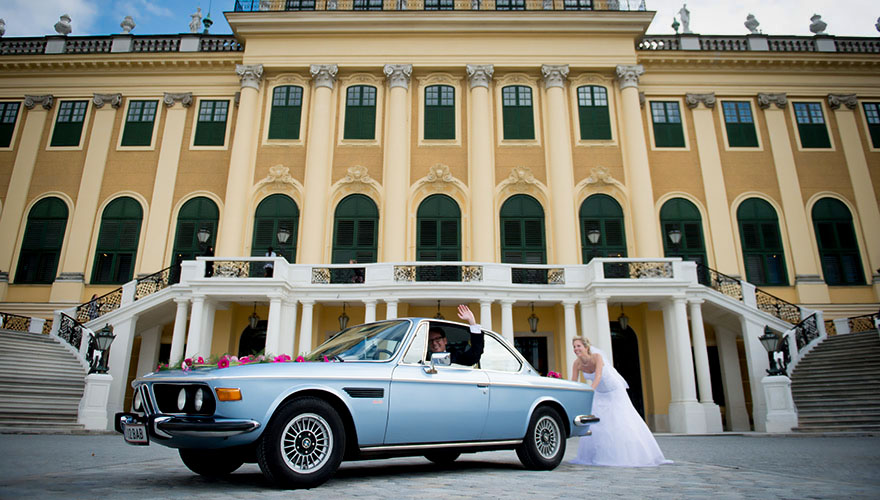 Vor dem Schloss Schönbrunn in Wien steht ein altes Auto indem der Bräutigam sitzt. Es scheint, als ob die Braut das Auto anschiebt.
