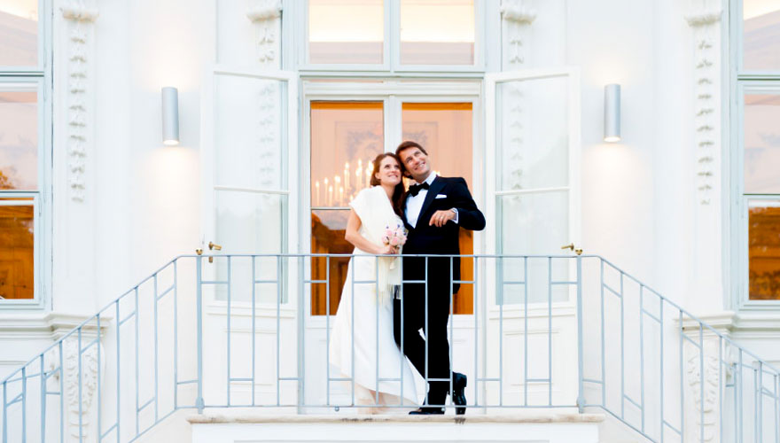Ein Brautpaar steht auf einem Treppenaufgang zu einem Tanzsaal. Hinter den Glasscheiben erkennt man den festlich beleuchteten Saal.