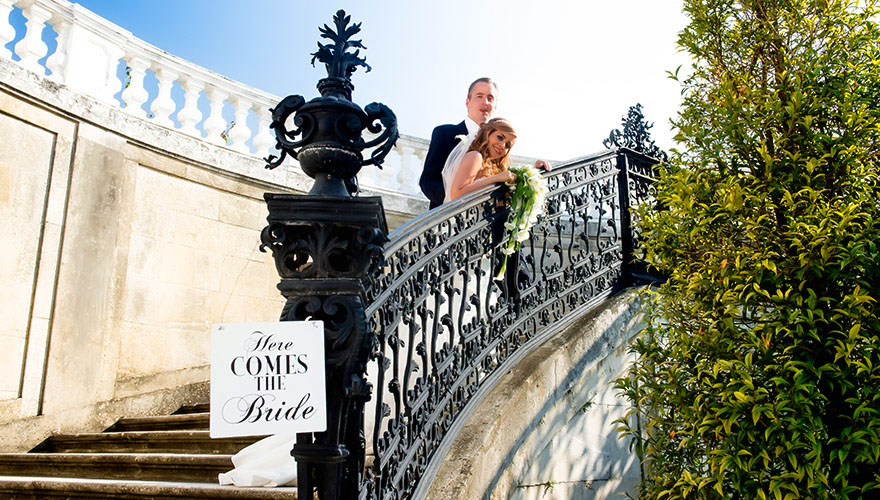 Auf einem Treppengeländer hängt ein Schild auf dem steht: Here comes the Bride. Weiter oben auf den Stufen steht das Brautpaar und blickt zum Fotografen.