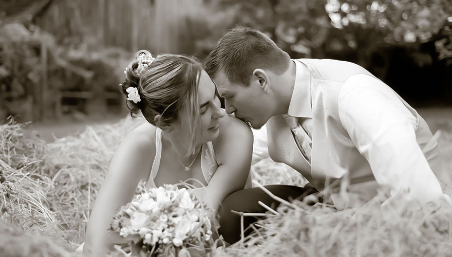 Vor einem Holzstadl ist ein Haufen Heu. In diesem Haufen liebkost ein Bräutigam seine Braut. Er küsst Ihre Schulter.