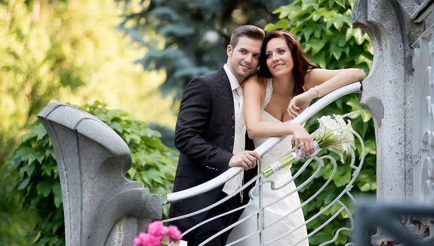 Ein Brautpaar steht auf einer Treppe und der Bräutigam umarmt seine Braut. Das Treppengeländer deutet auf ein älteres, gut erhaltenes, teures Haus hin.