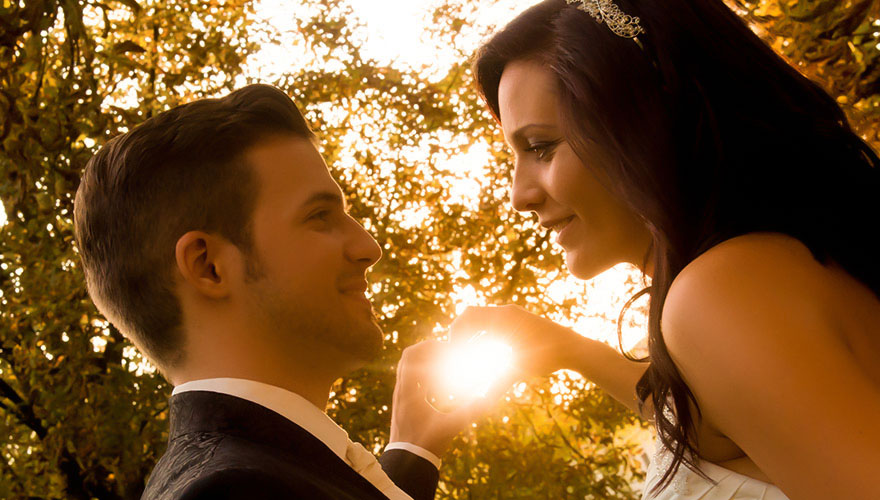 Ein Brautpaar steht unter Bäumen und geniesst die warmen Sonnenstrahlen welche sich ihren Weg durch die Blätter suchen. Beide lächeln.