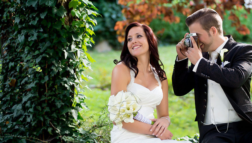 Braut und Bräutigam sitzen nebeneinander. Er fotografiert seine Braut, während sie sich wegdreht und lächelt. Das Bild wirkt herbstlich.