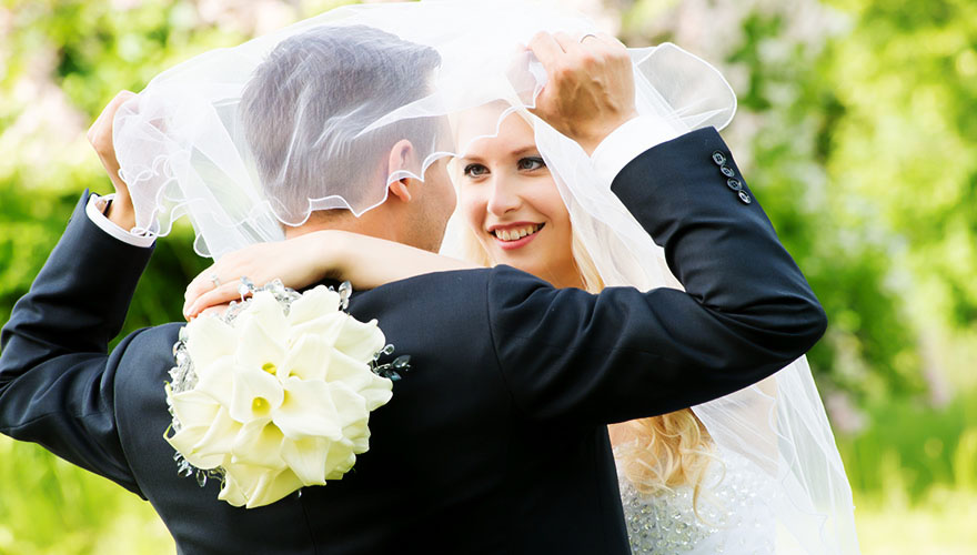 Ein Brautpaar steht ganz nah beieinander und der Bräutigam hat den Brautschleier über ihre Köpfe gelegt.