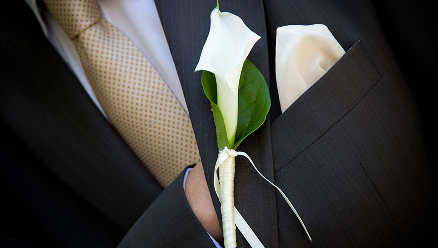 Ein Bräutigam steckt seine Hand in das Sakko. Auf der Weste hängt eine Callasblüte. In der Tasche steckt ein Tuch.