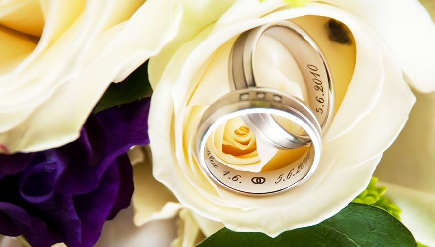 Auf einer Rosenblüte liegen zwei Eheringe. Die Rosenblüte ist Teil eines Blumenstrauss. In den Ringen steht ein Hochzeitsdatum.