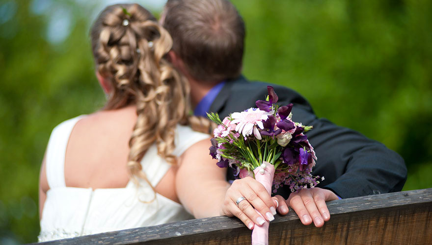 Ein Brautpaar sitzt auf einer Bank und schaut in den Himmel. Die Braut hat den Blumenstrauss nach hinten auf die Lehne gelegt und wir bemerken den Ring an ihrer Hand.