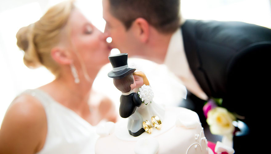 Ein detailierter Blick auf die Brautpaar-Figuren auf der Hochzeitstorte, welche sich küssen. Dahinter ist das echte Brautpaar und küsst sich auch.