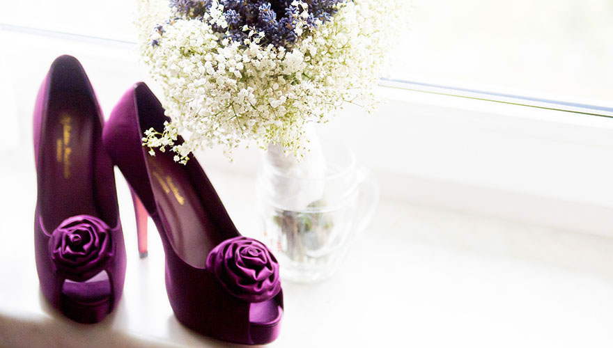 Auf einer Fensterbank stehen Brautschuhe mit hohen Stöckeln und Rosenaufsätzen auf der Oberseite der Schuhe. Dahinter steht ein Blumenstrauss mit Lavendel und Zierpflanzen.