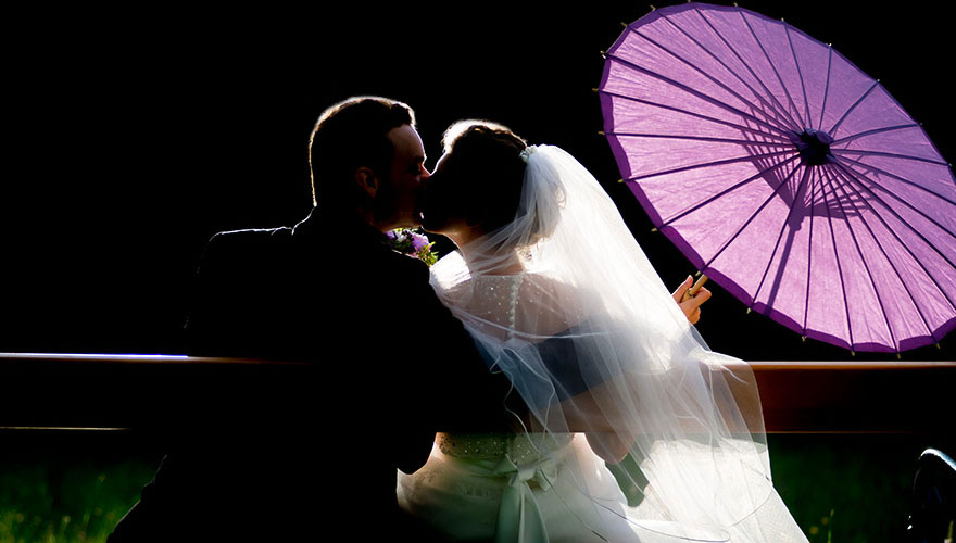 Ein Brautpaar sitzt in der Nacht auf einer Parkpank. Die Braut hält einen Sonnenschirm auf Papier in Ihrer rechten Hand, während sie den Bräutigam küsst.