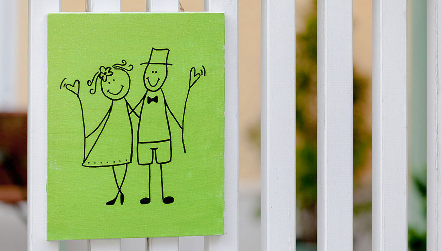 Auf einem Lattenzaun hängt ein Schild auf dem ein Brautpaar aufgezeichnet ist. Sie halten beide ein Herz in der Hand.