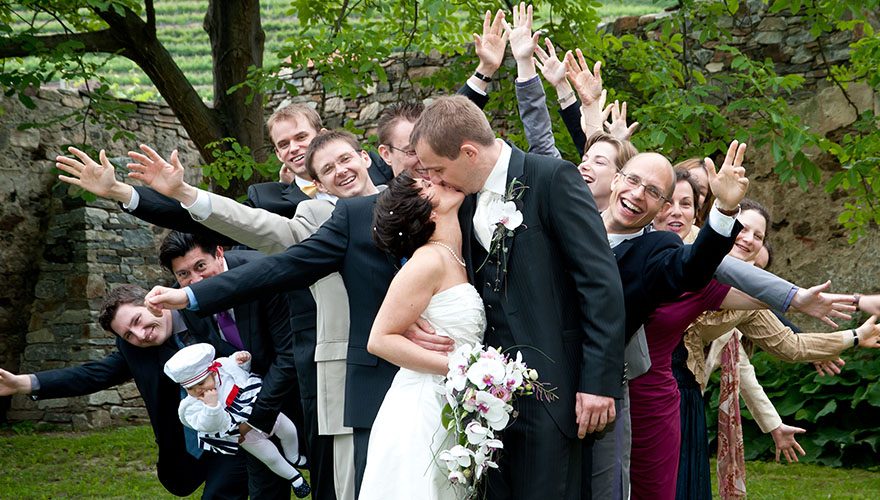 Ein Brautpaar küsst sich in einem Burgpark. Ganz dicht gedrängt stehen dahinter zwölf Hochzeitsgäste und winken dem Fotografen zu, schreien, lachen und freuen sich.