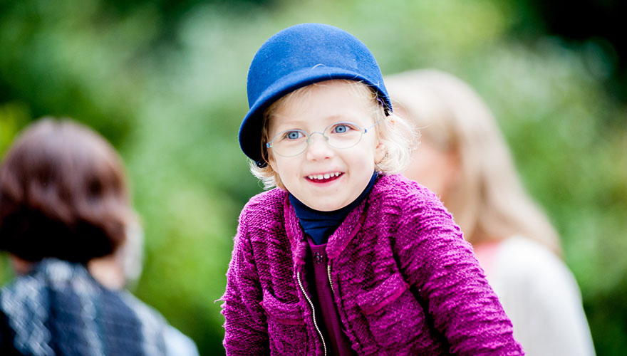 Ein Mädchen von ca. 4 Jahren mit einem modischen Hut und einer Brille lächelt jemanden zu.