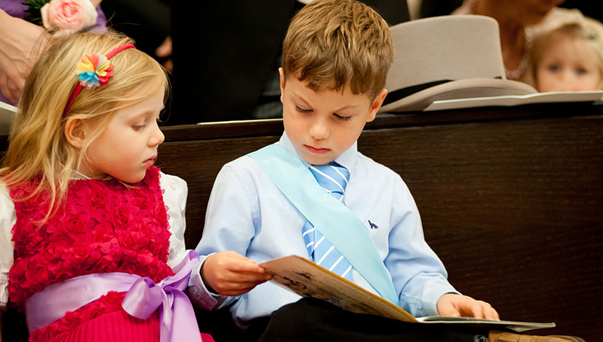 Auf einer Kirchenbank sitzen zwei Kinder, ein Mädchen und ein Knabe, während einer Hochzeit. Der Knabe liest in einem Bilderbuch und wird dabei von dem Mädchen beobachtet. Der Knabe trägt eine Krawatte und eine Scherpe. Im Hintergrund liegt ein Zylinde