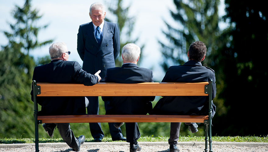 Bei einer Hochzeit. Drei Männer in Anzügen sitzen auf einer Parkpank. Ein Mann steht vor Ihnen und hört sich an, was ein sitzender Mann zu sagen hat.