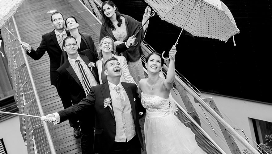 Ein Brautpaar und fünf Hochzeitsgäse tragen Schrime, denn es regnet. Sie geben kurz die Schirme beiseite und rufen zum Himmel, lachen und werden leicht nass.