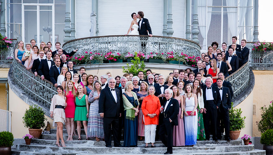 Eine Hochzeitsgesellschaft hat sich zu einem Gruppenfoto auf der festlichen Ausenstiege eines Palais eingefunden. Das Brautpaar steht am oberen Balkon und der Bräutigam küsst die Braut. Alle lächeln.