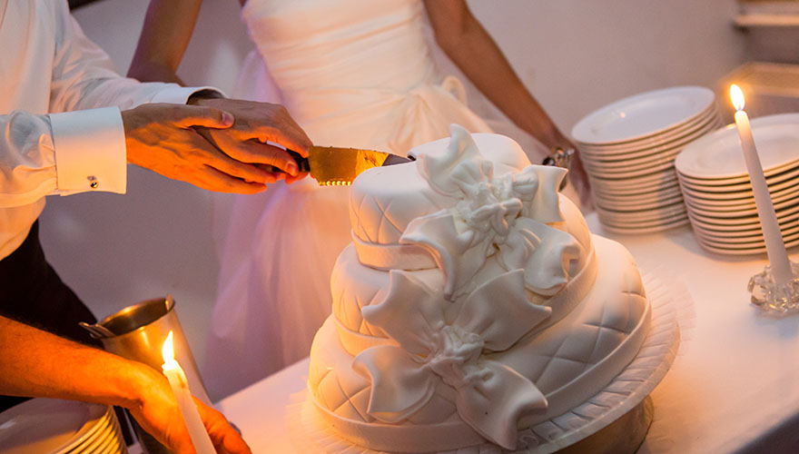 Ein Brautpaar schneidet eine Torte an. Die Torte ist mit Bändern und Maschen aus Zuckerguss verziert. Neben der Torte steht eine Kerze und viele Teller.