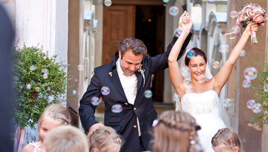 Ein Brautpaar schreitet aus der Kirche und wird von seinen Gästen jubelnd empfangen. Seifenblasen fliegen durch die Luft. Die Braut hebt die Hände und Lacht. Der Bräutigam hält die Braut an der Hand, seine Hand wird mitgerissen und er lächelt verstohl