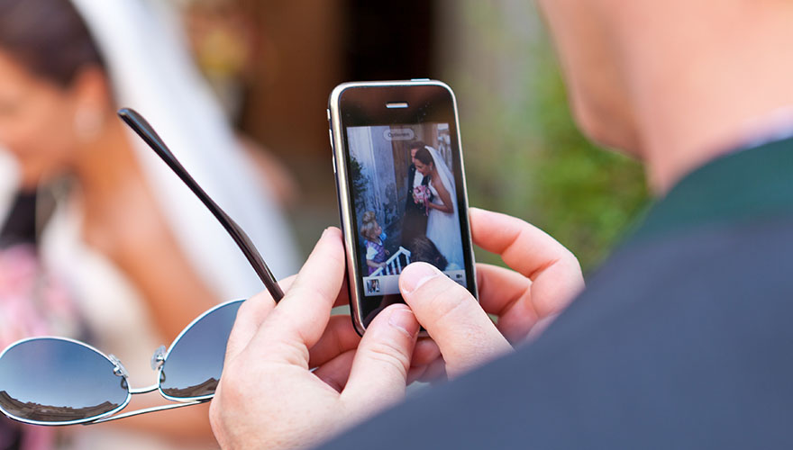 Bei einer Hochzeit hat ein Hochzeitsgast mit seinem Mobiltelefon ein Foto vom Brautpaar während der Gratulation eines Kindes gemacht. Er betrachtet das Foto. In der anderen Hand hält er seine Sonnenbrille, im Hintergrund erkennt man die Braut.