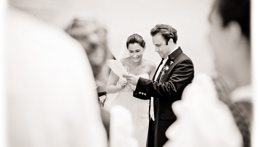 Ein Brautpaar liest eine Nachricht auf einem Blatt Papier und schmunzelt. Die Gäste sehen dabei zu. Der Betrachter ist mitten unter den Gästen.