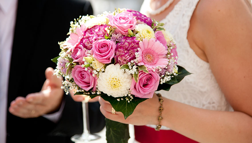 Ein Brautstrauss mit verschiednen Blüten, vor allem Rosen. Im Hintergrund erkennt man den Bräutigam der eine erklärende Geste miut der Hand macht.