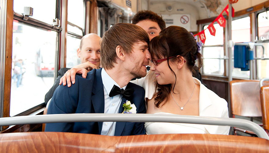 Ein frisch vermähltes Brautpaar sitzt in der Strassenbahn. Sie pressen die Stirn aneinander. Er flüstert ihr etwas zu. Hinter ihnen sitzen zwei Männer und lächeln während sie das Brautpaar beobachten.