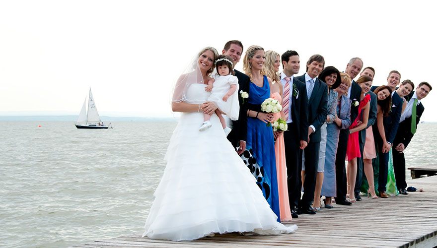 Ein Brautpaar und deren kleine Tochter stehen vor einigen Hochzeitsgästen an einem Steg. Im See fährt ein kleines Segelboot. Einge Gäste versuchen an den anderen vorbeizusehen um auch am Foto zu sein.