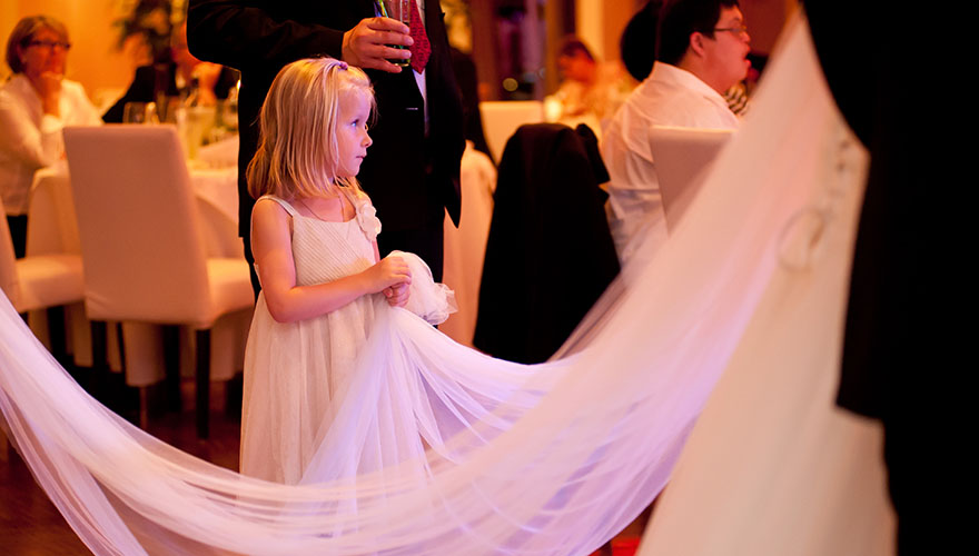 Eine Braut schreitet durch die Hochzeitsgäste, welche bei der Hochzeitstafel bereits sitzen. Ein kleines Mädchen hält dabei den langen Brautschleier damit er nicht den Boden berührt.