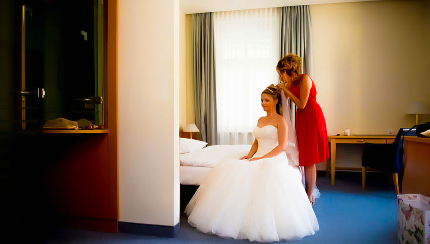 In einem Hotelzimmer sitz eine Braut fertig angezogen. Eine Freundin steckt ihr den Brautstrauss auf die Haare.