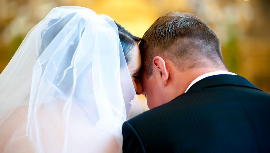 Ein Brautpaar sitzt vor dem Traualtar und die Braut flüstert ihrem Bräutigam etwas ins Ohr.