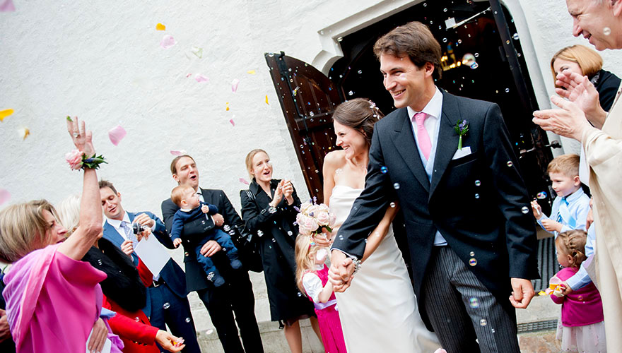 Ein Brautpaar kommt Hand in Hand aus der Kirche und wird von Ihren Hochzeitsgästen mit Seifenblasen und Rosenblüten, welche in die Luft geworfen werden, begrüsst. Die Braut weicht lachend etwas aus. Der Priester klatscht.