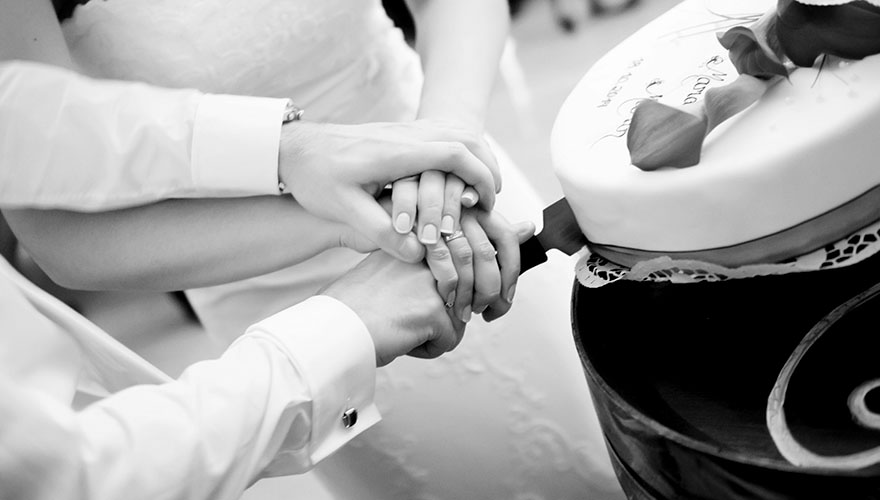 Ein Brautpaar schneidet eine Torte an. Auf dem Messergriff liegt erst die Hand des Bräutigams und dann die Hand der Braut. Es folgt die zweite Hand der Braut und dann die zweite Hand des Bräutigams.