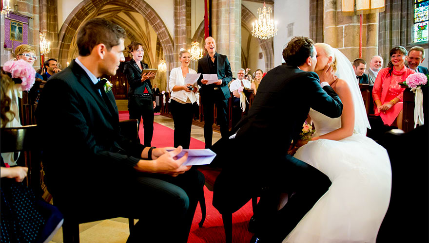 Ein Hochzeitspaar wird mit einer Gesangsdarbietung in der Kirche überrascht. Die drei Sänger stehen im Mittelgang. Vorne küsst der Bräutigam seine Braut. Die Gäste folgen dem Geschehen entzückt.