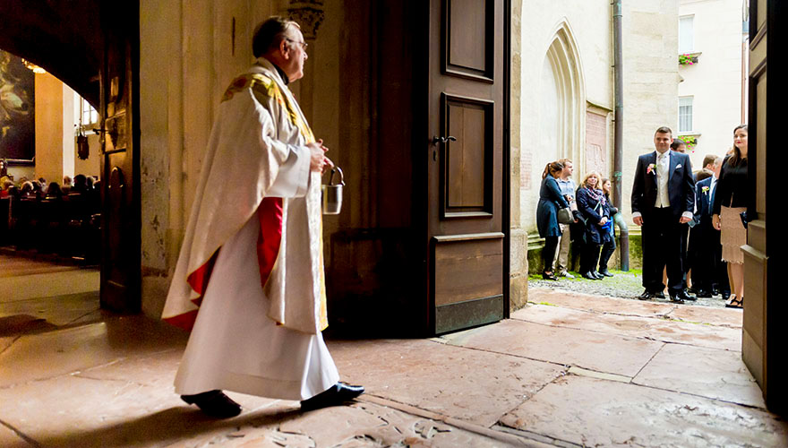 Ein Priester schreitet von der Kirche ins freie um einen Bräutigam und seine Gäste zur Trauung zu führen.