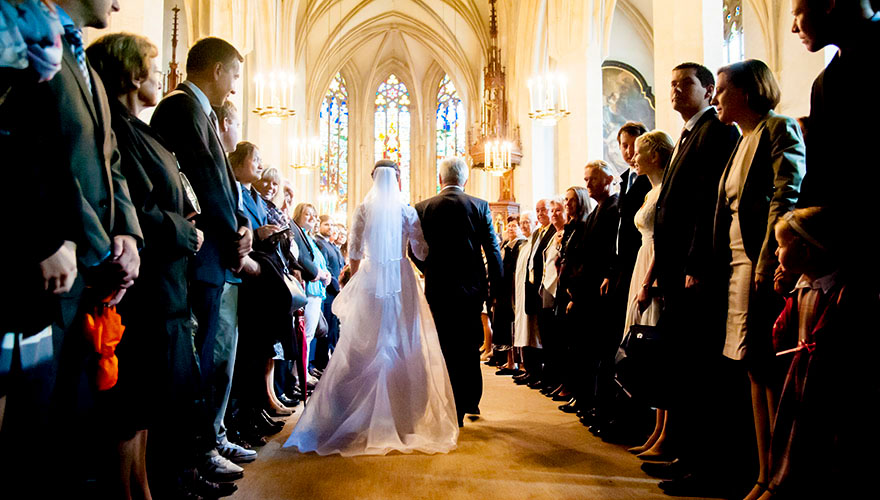Eine Braut und Ihr Vater schreiten durch den Mittelgang einer Kirche zur Trauung. Die Gäste stehen und bewundern die Braut.